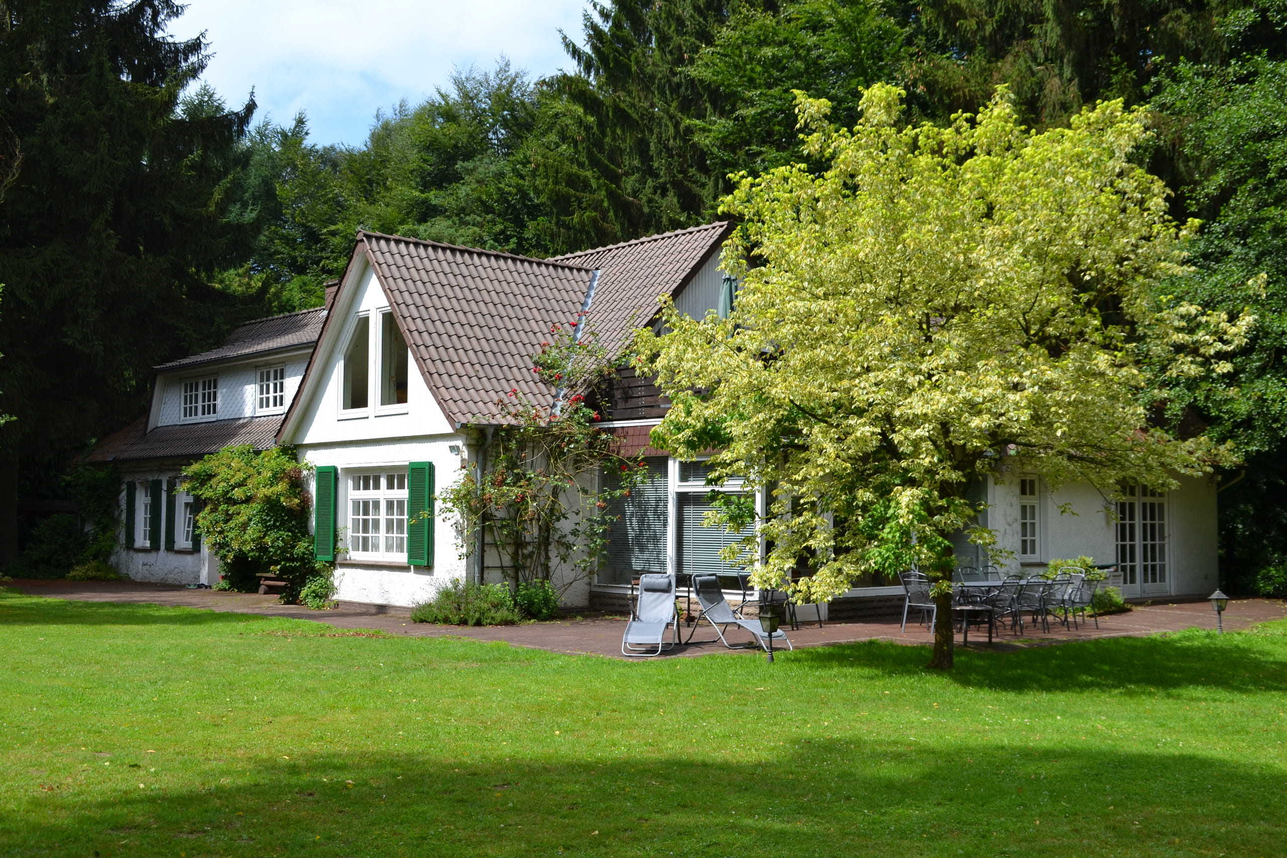 Haus mit grünen Fensterläden und großer Terrasse, davor grüner Rasen, dahinter Wald